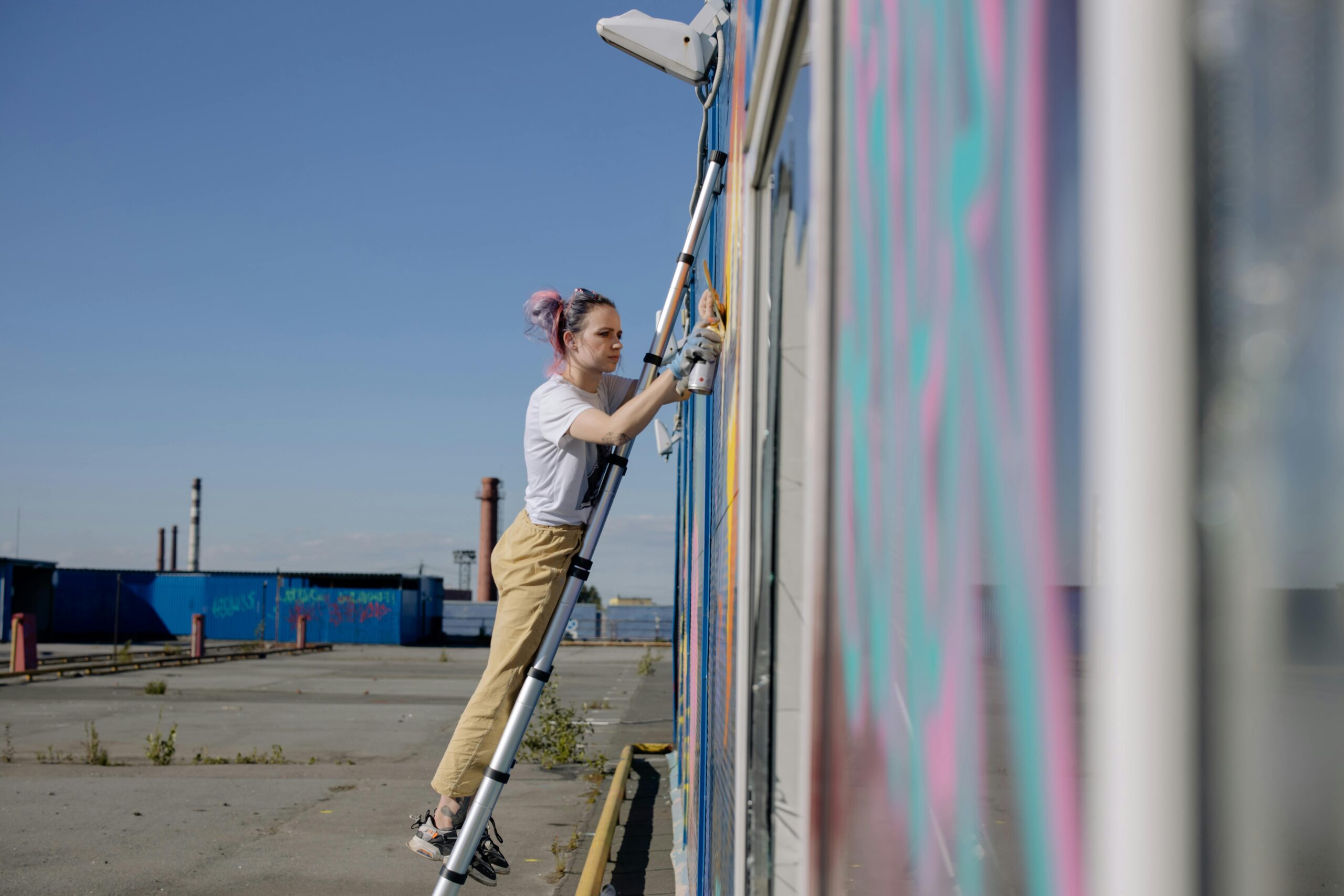 A female street artist on a ladder creating a vibrant mural outdoors.